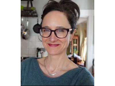 Mary Purdy smiling, wearing red lipstick, black glasses, a silver necklace, and green shirt. Her dark brown hair is pulled back into a bun.