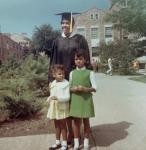 LaRae Kemp wearing a black cap and gown, standing next to her two daughters. One wearing a white sweater and yellow dress, and the other wearing a green dress.