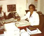 LaRae Kemp wearing a white doctor's coat sitting at her office desk. On the desk are books, strewn papers, a phone, a flower vase, and coffee mug.