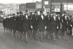 A group of women in uniform suits and hats saluting with their right hands, marching in formation down a city street.