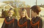 Maureen Garrity with two women in dark military-style uniforms with orange braided cords and hats stand outdoors in front of a white house and a brick building.