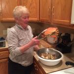 Maureen wearing a gray long-sleeved shirt, stands in a kitchen holding a cooked crab with tongs. On the counter beside her is a white bowl filled with crab legs.