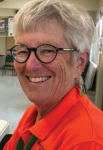 Smiling woman with short gray hair and blue rimmed glasses wearing an orange shirt.