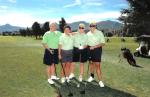 Four people standing on a golf course with mountains in the background, holding golf clubs and wearing green shirts and dark shorts.