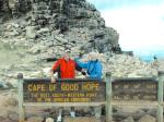 Maureen wearing a blue turtleneck, with Nan Ryan, wearing a red shirt and hat, standing behind a wooden sign at the Cape of Good Hope; rocky terrain, cliffs and the ocean are in the background.
