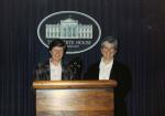 Maureen Garrity and Judith McCaan, both wearing button down shirts and blazers, stand behind a podium with microphones; a dark curtain behind them displays the White House emblem and the text "THE WHITE HOUSE WASHINGTON."