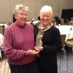 Maureen and Nan standing together in a room with round tables and chairs. Nan is wearing a pink turtleneck and black pants. Maureen is wearing a black suit with a gold metallic cowl neck shirt and holding a glass award.