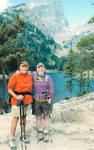 Maureen wearing a purple pullover, olive green shorts and a hat, with Nan Ryan, wearing an orange pullover, khaki shorts and a hat, posing in front of a scenic mountain lake surrounded by pine trees.