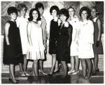 Nine women wearing formal dresses, arranged in two rows in front of a wall with diagonal wooden panels.