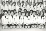 A group of women in three rows, the front two rows are kneeling and the third row is standing All women are wearing dark bottoms, tops, and white blazers with a chest logo.