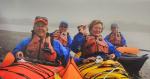 Five people wearing blue jackets and red life vests sit in brightly colored kayaks—one orange and one yellow—on a body of water. The background is foggy.