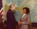 LaRae Kemp wearing a brown suit shaking hands with a man in military uniform while standing in front of a large world map. An American flag is visible on the left side.