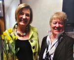 Terri Blevins, left, wearing a bright green jacket with large buttons and a chunky beaded necklace holding a bunch of yellow tulips. Maureen, right, wears a dark blazer over a white shirt with a scarf.