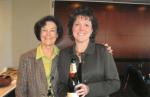 Mary Krugman in a brown plaid jacket stands with Carolyn Sanders, who wears a khaki shirt and holds a bottle of Moet champagne.