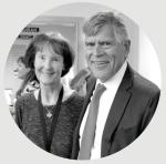 Black-and-white photo of a smiling woman in a dark top with a cross pendant and short hair standing beside a man in a suit and tie.