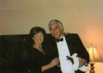 Smiling dark-haired woman in a black dress with pearls holds a glass, arm around a man in a tuxedo holding champagne wrapped in a white cloth.