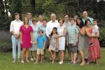 Extended Krugman family, including Mary and Dick, with children and grandchildren in casual attire standing together on a grassy lawn with trees.