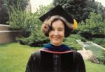 Smiling dark-haired woman in a black PhD cap and gown standing on grass with a brick wall and garden behind her.