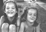 Black-and-white photo of two young girls sitting outdoors; one grins gap-toothed, the other smiles softly with eyes closed.