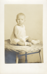 A black and white portrait of a baby in a white cotton sleeveless dress wearing white socks and shoes sitting on a round tan colored wicker stool.