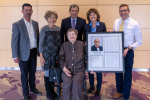 Three men and two women wearing business attire, smiling, and standing around Bonnie Camp, MD who is seated, wearing a brown pantsuit and holding a framed portrait of Stephen Berman, MD.
