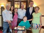 Three men and three women smiling and standing around Bonnie Camp, MD who is seated, wearing a teal shirt and an oxygen cannula. A framed award with a gold ribbon is sitting on a table in front of Dr. Camp.