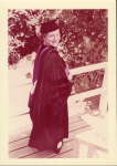 Bonnie Camp wearing a black graduation cap and gown standing on a wooden deck next to a railing with leafy trees in the background.