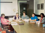 Five people in a meeting, circa late 1980's, seated around a long conference table and posing informally for the camera.