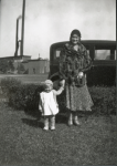 Black-and-white photo of a woman in a fur coat and patterned dress standing beside a young child in a light-colored coat. They hold hands in front of a 1940's car.