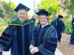 Two older women wearing doctoral graduation regalia and mortarboards stand together outdoors on a tree-lined university campus.
