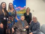Four women smiling and standing around Bonnie Camp, MD who is seated and wearing a brown tunic. A vibrantly colored painting of mountains hangs on the wall behind the women.