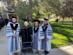 Four women wearing doctoral graduation regalia and mortarboards stand together outdoors on a tree-lined university campus.