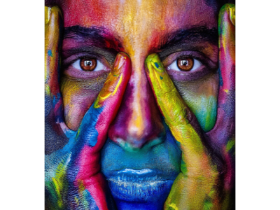 Photograph close-up of a woman's face, holding two fingers on either side of the bridge of the nose. The person's eyes are brown and their skin has been painted with multiple bright colors, including red, pink, blue, purple, yellow, green, and orange.
