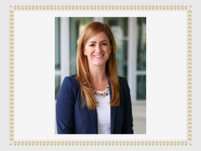 Kelsey Schwander. Smiling woman with long straight red hair wearing a navy blue blazer, white blouse and multi colored necklace.