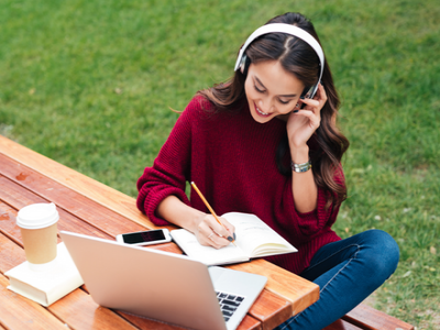 Young woman sitting at a table outside studying with headphones, a laptop, and a pen and notebook.