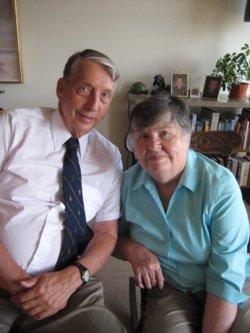 An elderly couple sitting close together indoors. The man, wearing a light pink shirt and a dark tie, and the woman, wearing a light blue blouse, both smile gently. Behind them is a bookshelf with books, family photos, and a potted plant.