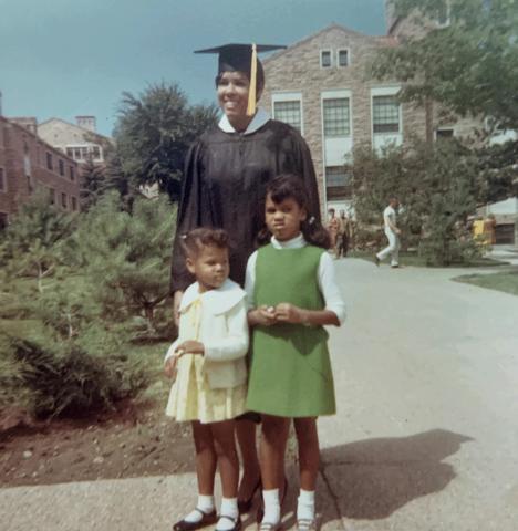 LaRae Kemp wearing a black cap and gown, standing next to her two daughters. One wearing a white sweater and yellow dress, and the other wearing a green dress.