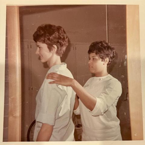 During an exercise for their physical therapy program, a female student with short brown hair stands with her back to the camera while LaRae Kemp in a white uniform, places her hands on the student's shoulders.