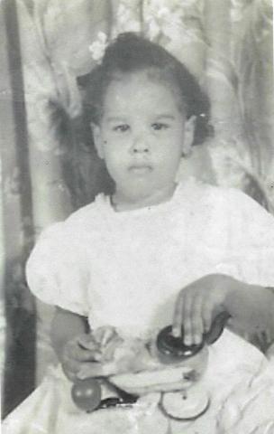 LaRae at age 2 seated in front of a floral curtain backdrop wearing a white dress, holding a toy. 