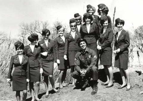 Nine women and one man in military uniforms; women stand on a hill while the man crouches in front. Leafless trees and a clear sky in the background.