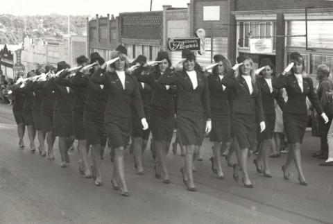 A group of women in uniform suits and hats saluting with their right hands, marching in formation down a city street.