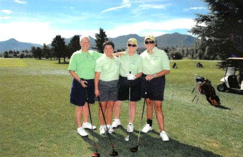 Four people standing on a golf course with mountains in the background, holding golf clubs and wearing green shirts and dark shorts.