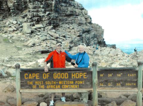 Maureen wearing a blue turtleneck, with Nan Ryan, wearing a red shirt and hat, standing behind a wooden sign at the Cape of Good Hope; rocky terrain, cliffs and the ocean are in the background.