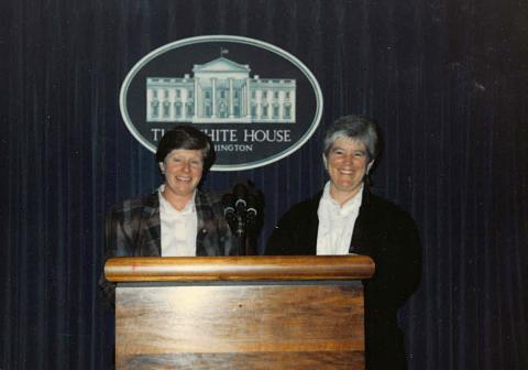 Maureen Garrity and Judith McCaan, both wearing button down shirts and blazers, stand behind a podium with microphones; a dark curtain behind them displays the White House emblem and the text "THE WHITE HOUSE WASHINGTON."
