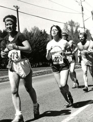 Maureen Garrity wearing shorts, a T-shirt and bib number 973 in a road race; three other runners, trees and power lines are in the background.