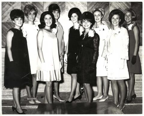 Nine women wearing formal dresses, arranged in two rows in front of a wall with diagonal wooden panels.