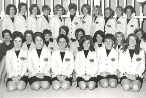 A group of women in three rows, the front two rows are kneeling and the third row is standing All women are wearing dark bottoms, tops, and white blazers with a chest logo.