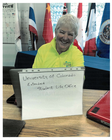 Maureen Garrity in a yellow polo shirt sits at a desk with a laptop labeled "University of Colorado Extended Student Life Office".