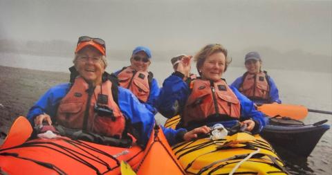 Five people wearing blue jackets and red life vests sit in brightly colored kayaks—one orange and one yellow—on a body of water. The background is foggy.