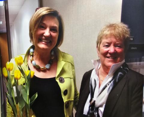 Terri Blevins, left, wearing a bright green jacket with large buttons and a chunky beaded necklace holding a bunch of yellow tulips. Maureen, right, wears a dark blazer over a white shirt with a scarf.